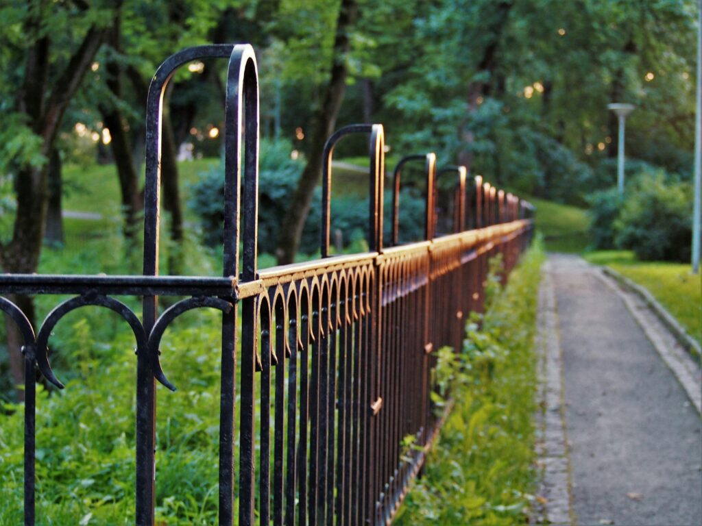 Sunset light glimmers on metal fence in lush park pathway, creating a serene outdoor ambiance.